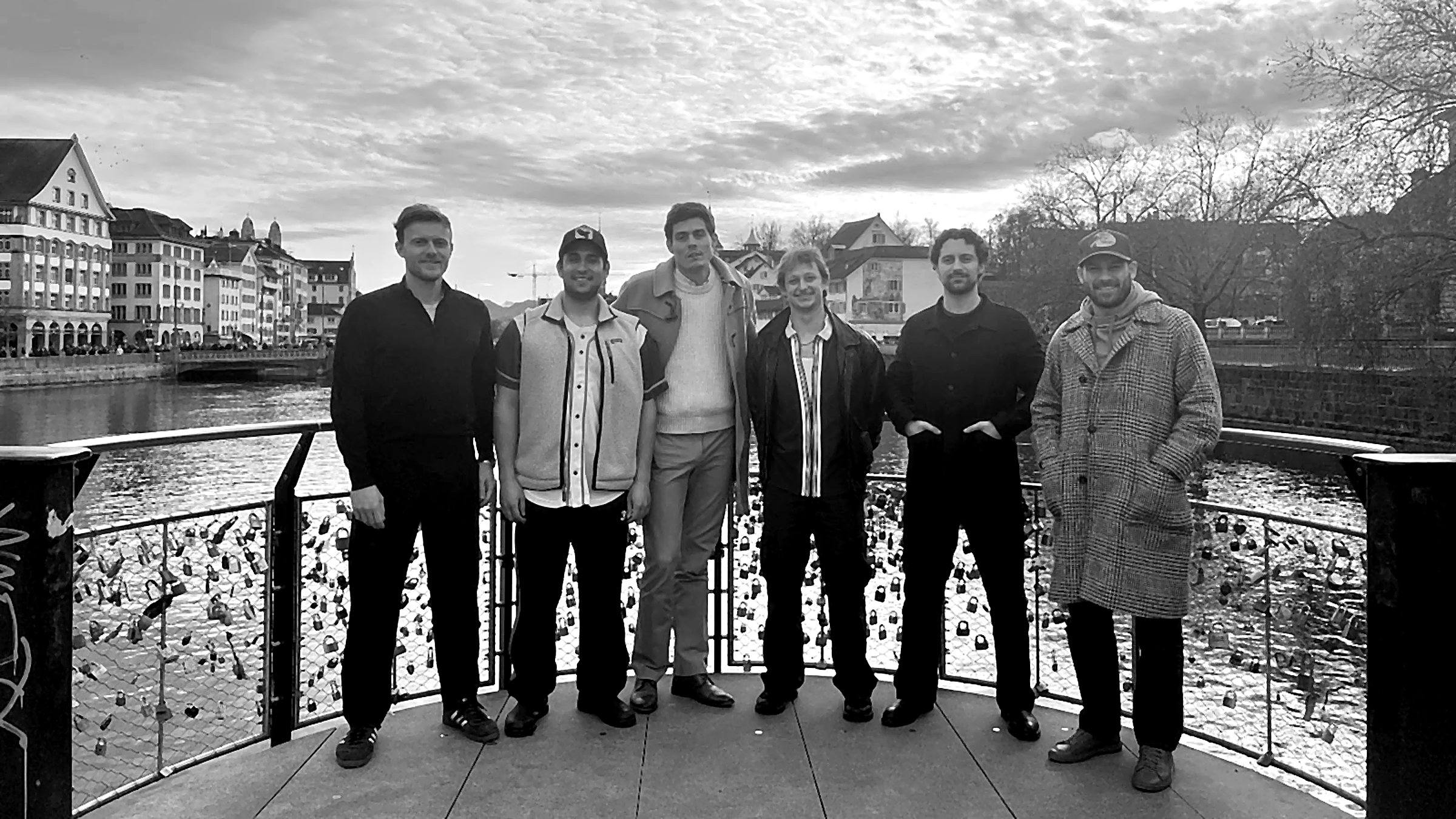 The six founders standing together on a viewing platform above the Limmat river in Zurich, under a cloudy sky.